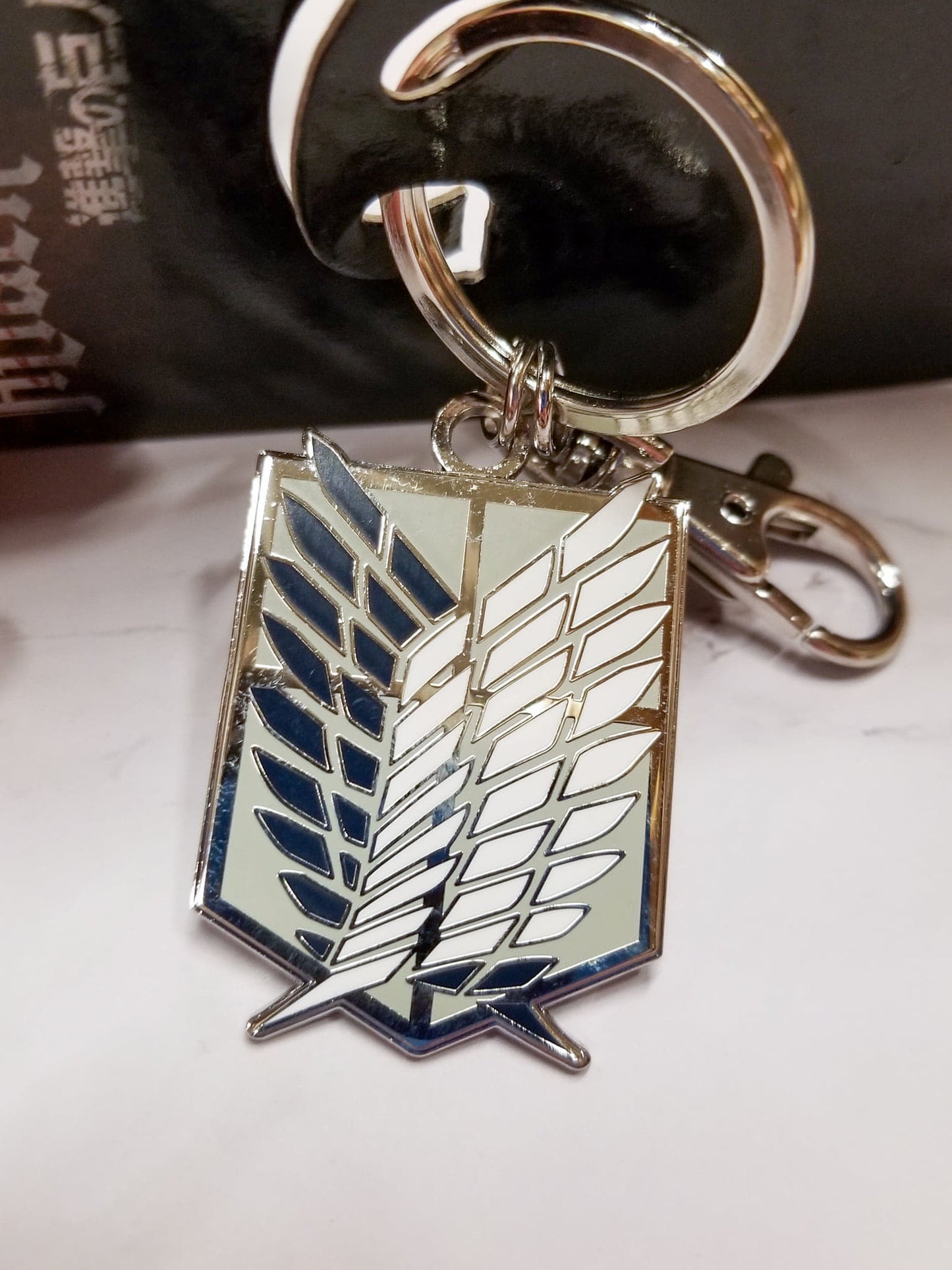 An enameled metal keychain with clip and ring, depicting the Scout Regiment (Scout Legion) emblem from Attack on Titan.  The metal emblem is approximately 1 inch wide by 1.5 inches tall.