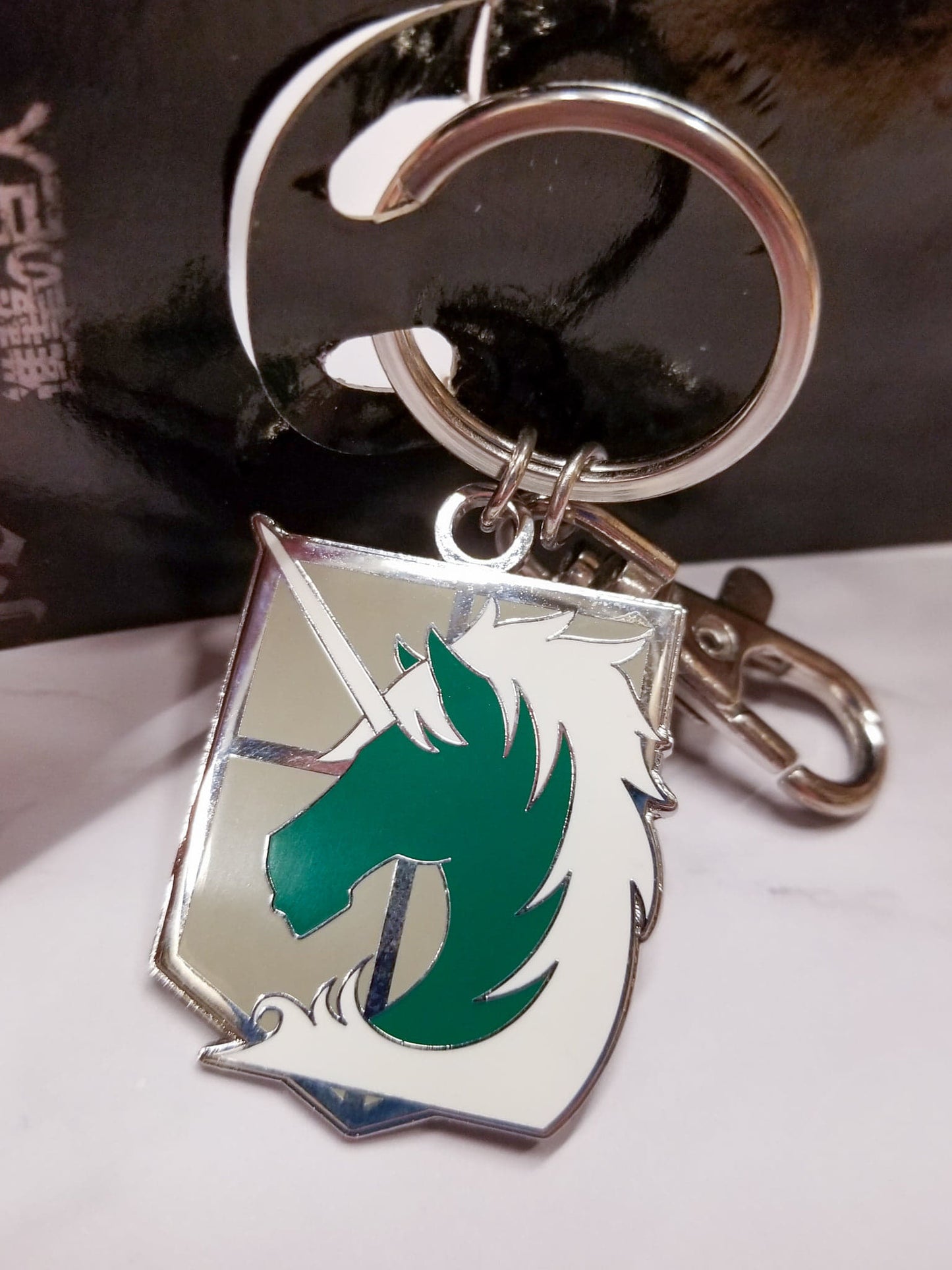 An enameled metal keychain with clip and ring, depicting the Military Police Brigade emblem from Attack on Titan.  The metal emblem is approximately 1 inch wide by 1.5 inches tall.
