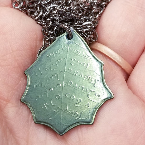 This Elvish Mithril Leaf of Spring Necklace is crafted in celebration of The Lord of the Rings by J. R. R. Tolkien. The leaf-shaped coin is struck from solid niobium, measures 2.7 cm in diameter, and weighs about 6.5 grams. Includes a 30" antique bronze cable chain with clasp. Coin artwork by Greg Franck-Weiby.  Coins are struck one at a time in the USA using antique machinery and traditional coining techniques. A colorful description is included with history, translations, and facts about the coins.